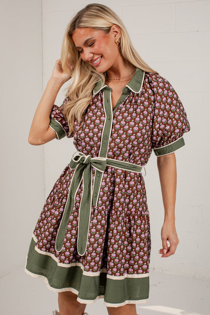 A woman modeling a short-sleeve button-front dress with a brown and pink floral print, olive green trim, and a matching olive green tie belt, set against a white brick wall.