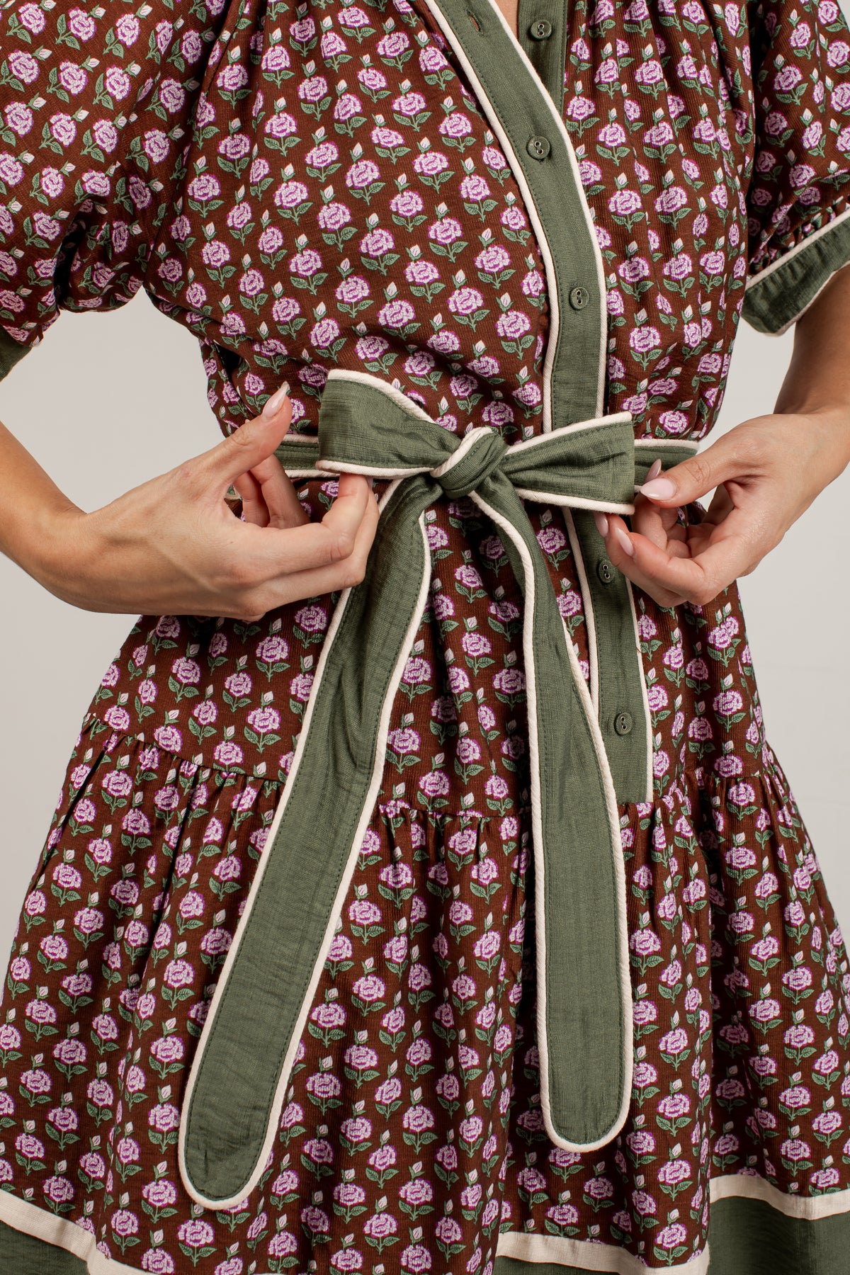 Close-up of a person wearing a brown dress with a pink floral print and an olive green self-tie belt with cream trim, adjusting the belt at the waist.