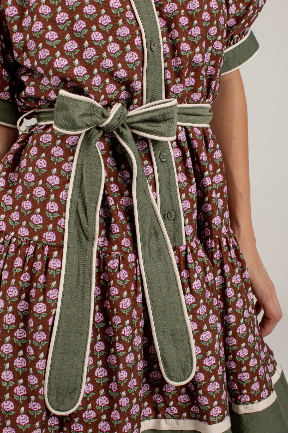 Close-up of a dress with a brown base, pink and green floral print, and an olive green self-tie belt with cream piping.