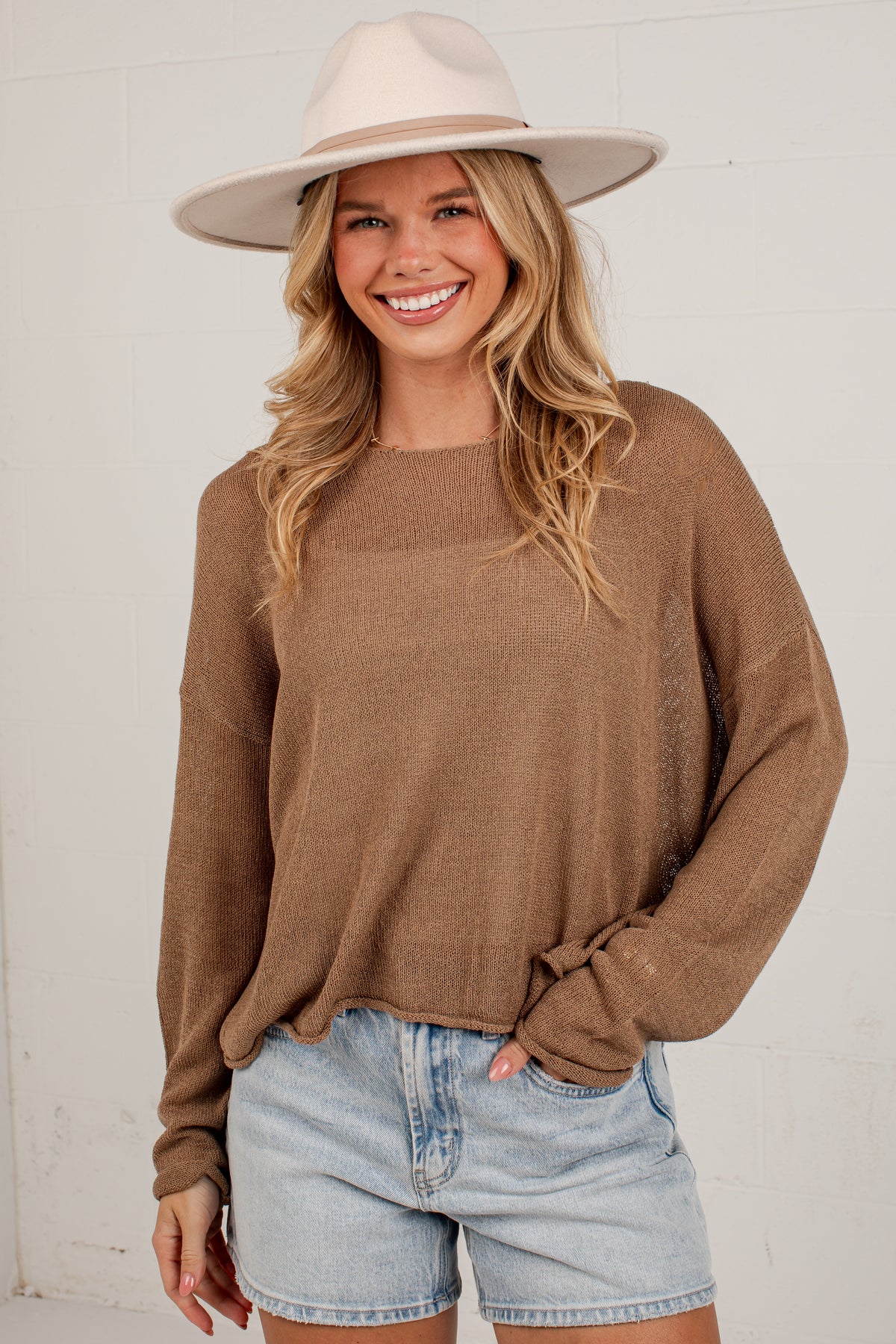 A smiling blonde woman wearing a brown knit long-sleeve sweater, light-wash denim shorts, and a beige wide-brim hat, standing against a white wall.