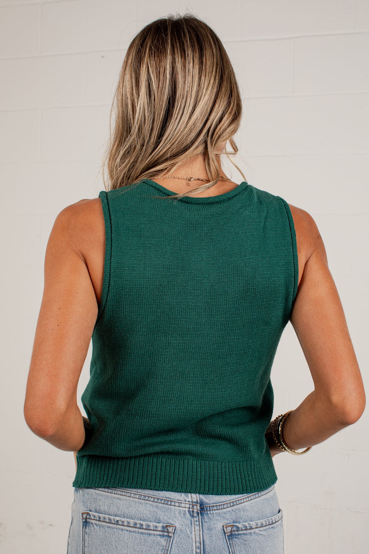 A woman viewed from behind wearing a sleeveless dark green knit top and light blue jeans against a white brick wall.