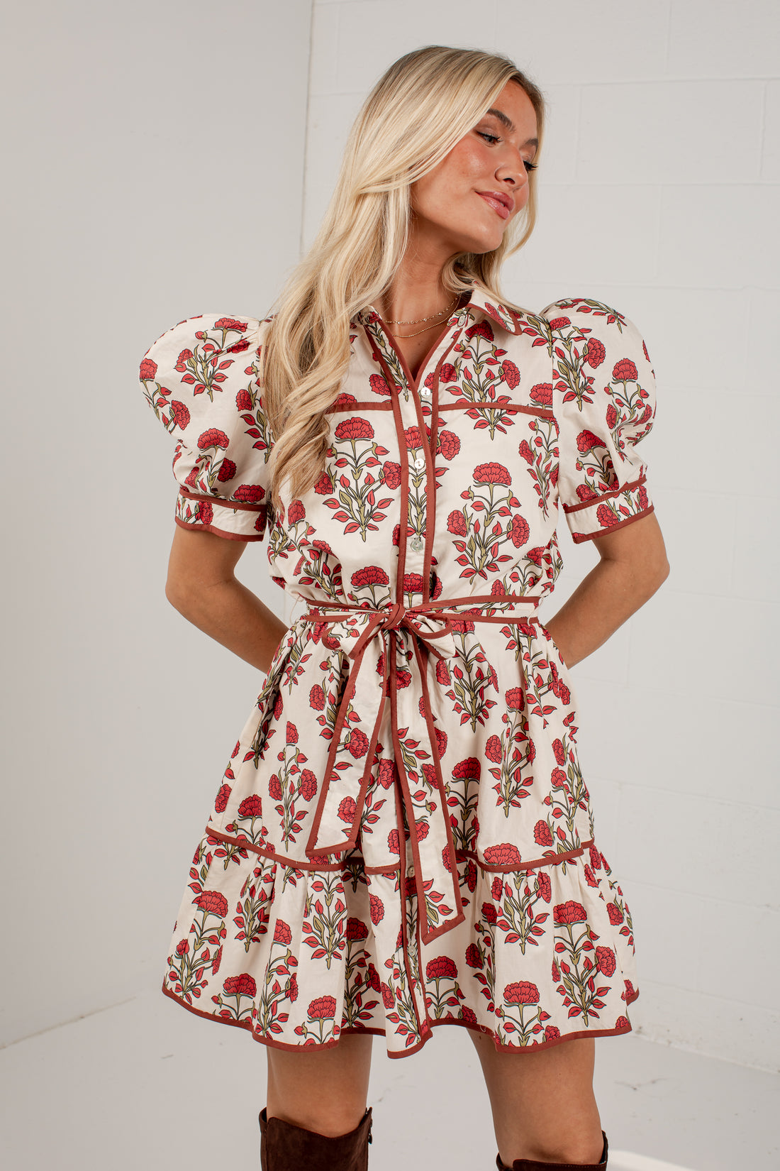 A woman modeling a short puff-sleeve dress with a red and green floral print on a cream background, featuring a tie waist belt and tiered skirt, set against a plain white studio background.