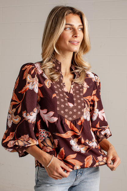 A woman wearing a brown floral blouse with puff sleeves and a gathered peplum hem, paired with light blue jeans, standing against a white brick wall.