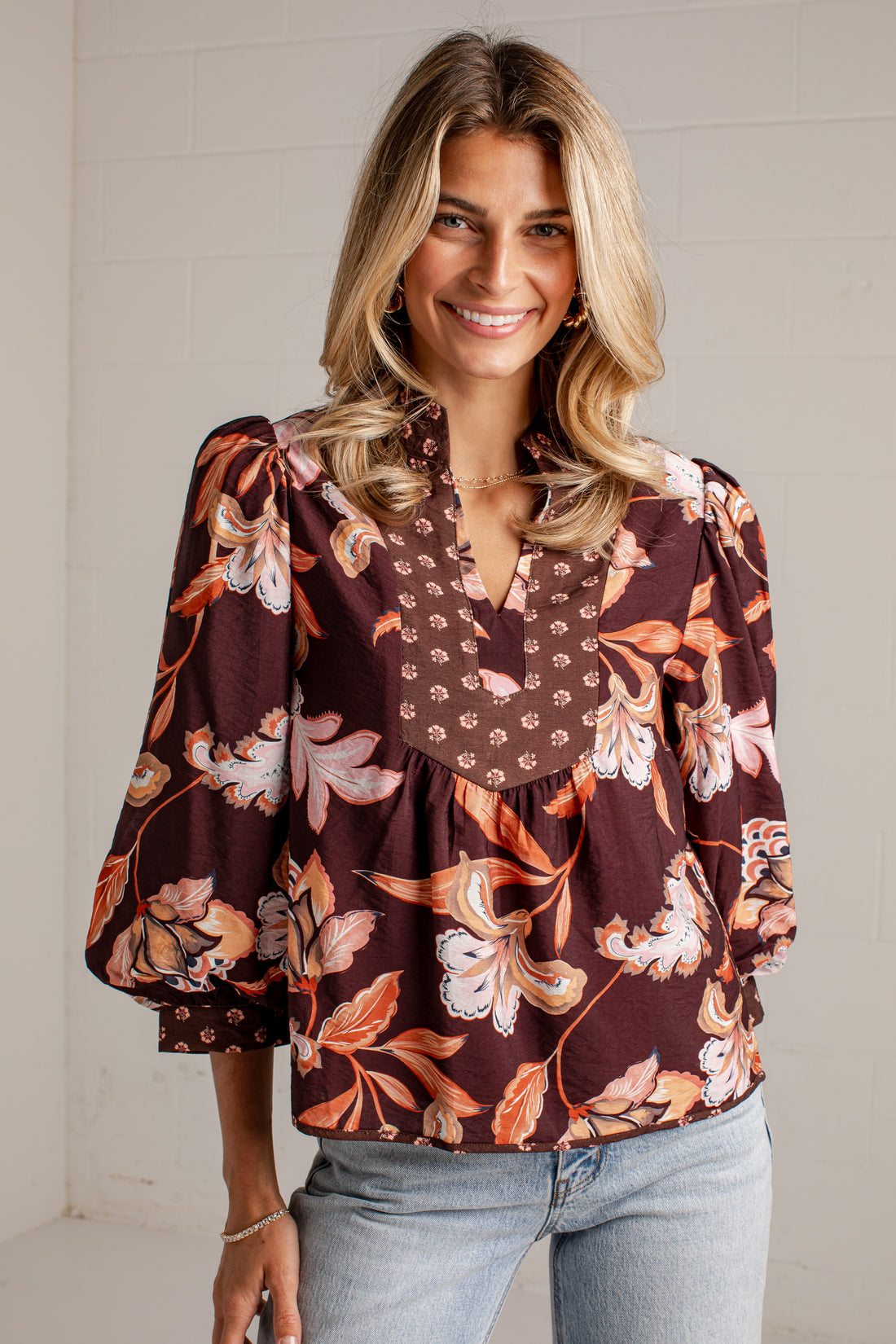A smiling blonde woman wearing a brown floral long-sleeve blouse with a V-neck and light-wash jeans, standing against a white brick wall.