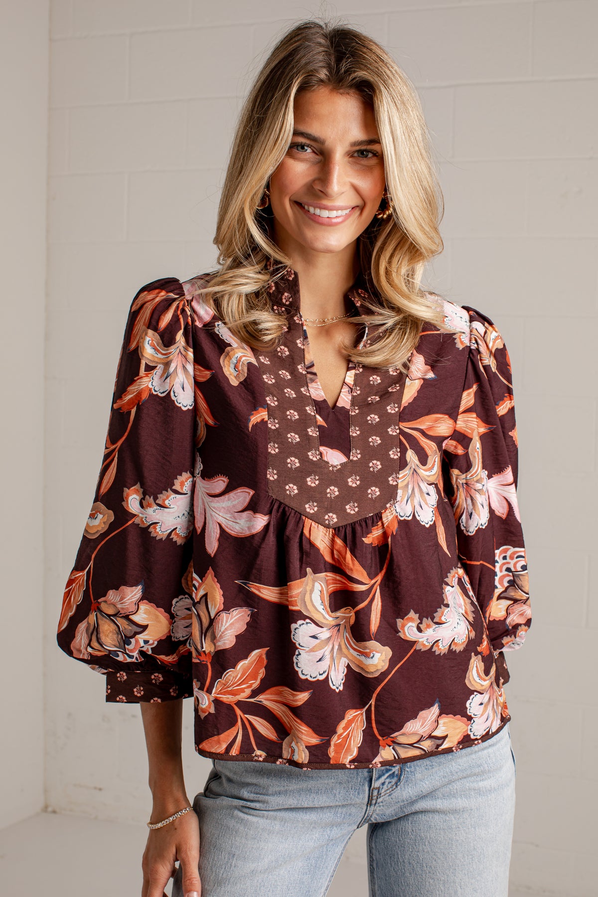 A smiling blonde woman wearing a brown floral long-sleeve blouse with a V-neck and light-wash jeans, standing against a white brick wall.