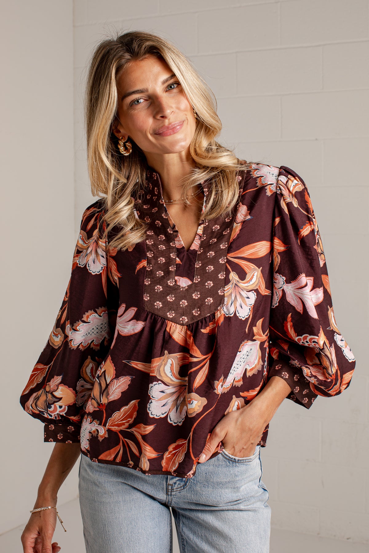 A woman wearing a brown floral blouse with puff sleeves and light blue jeans, standing against a white brick wall.