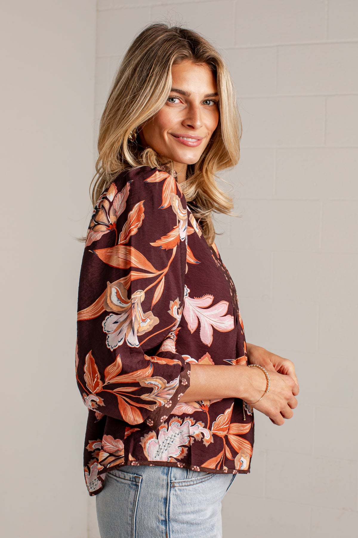 A woman wearing a dark brown floral blouse with orange, pink, and white botanical patterns, paired with light blue jeans, standing against a light gray brick wall.