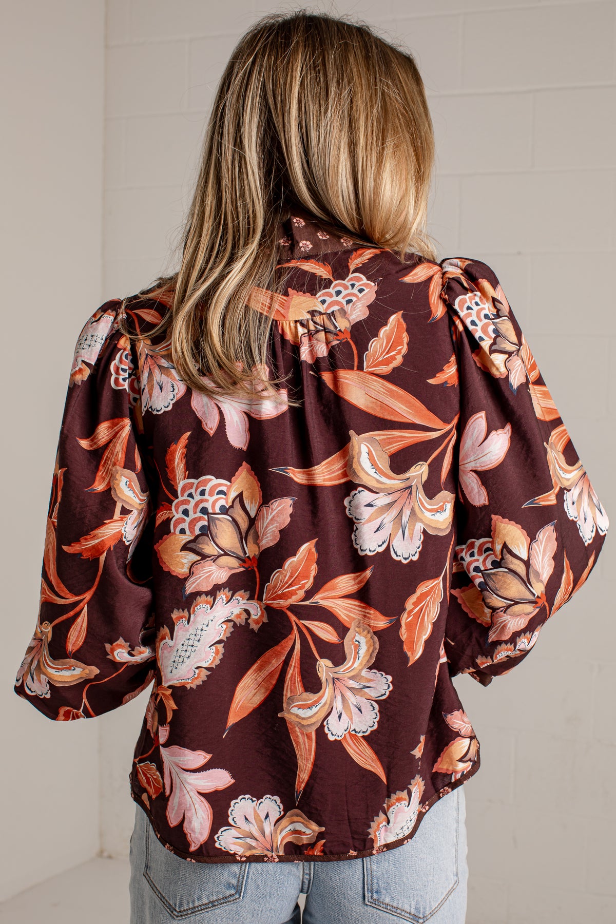 A woman viewed from behind wearing a dark brown floral blouse with puff sleeves and light blue jeans, standing against a white brick wall.