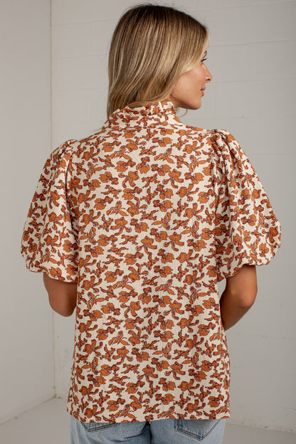A woman shown from the back wearing a short-sleeve blouse with puff sleeves and an allover brown floral print on a cream background, paired with light blue jeans against a white wall.