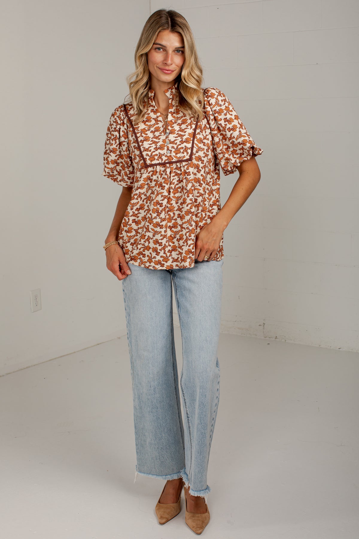 A woman modeling a short-sleeve button-up blouse with a rust orange and white floral pattern, paired with light-wash wide-leg jeans and tan pointed-toe heels, standing against a plain off-white studio wall.