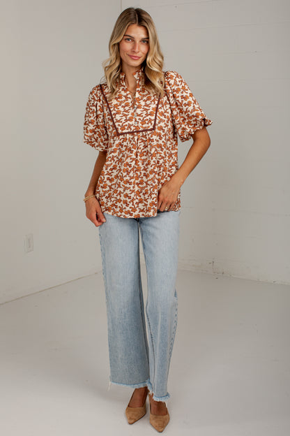 A woman modeling a short-sleeve button-up blouse with a rust orange and white floral pattern, paired with light-wash wide-leg jeans and tan pointed-toe heels, standing against a plain off-white studio wall.