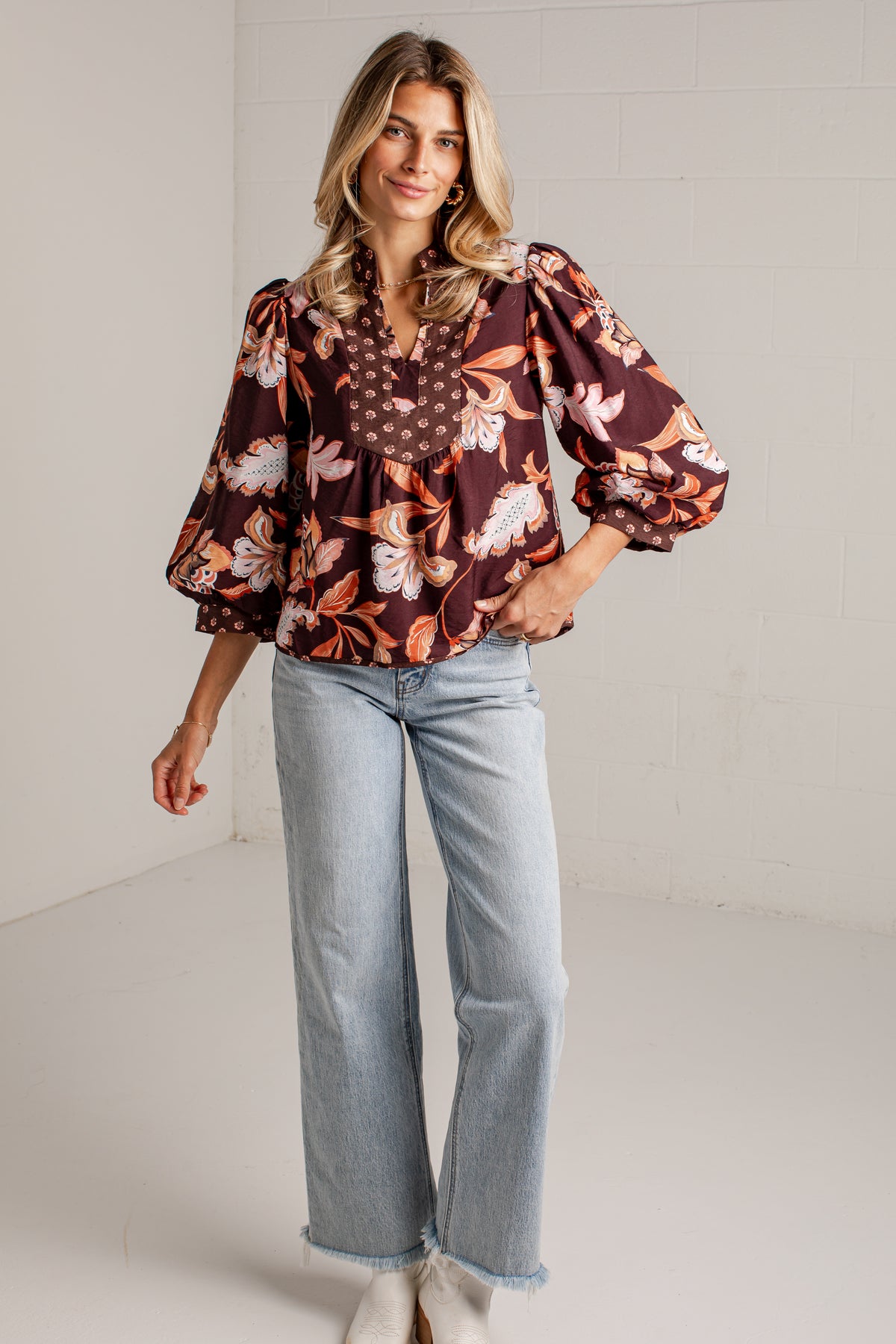 A woman modeling a brown floral blouse with puff sleeves and light-wash wide-leg jeans against a white studio wall.