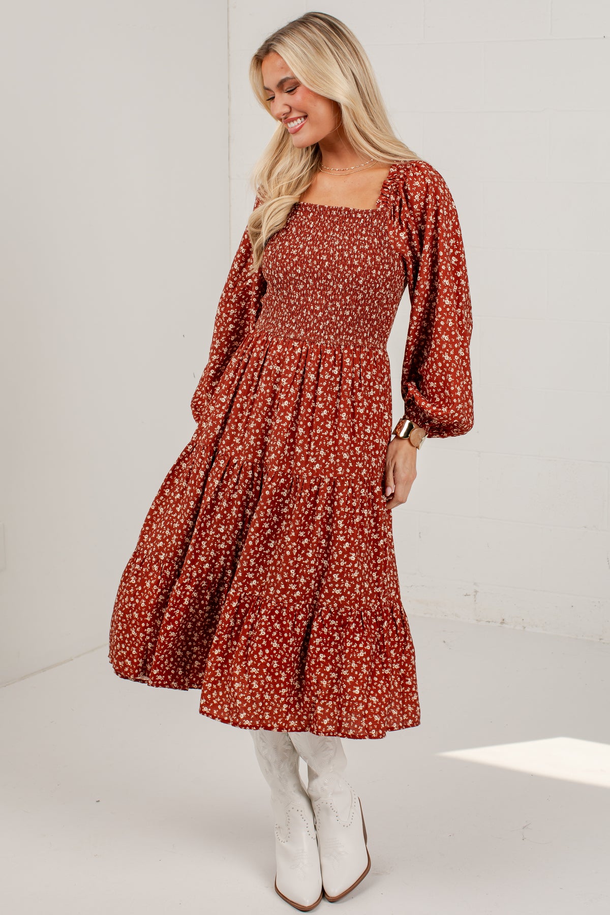 A woman modeling a rust-colored midi dress with a white floral print, featuring a smocked bodice, square neckline, and voluminous long sleeves, paired with white cowboy boots against a white brick wall background.
