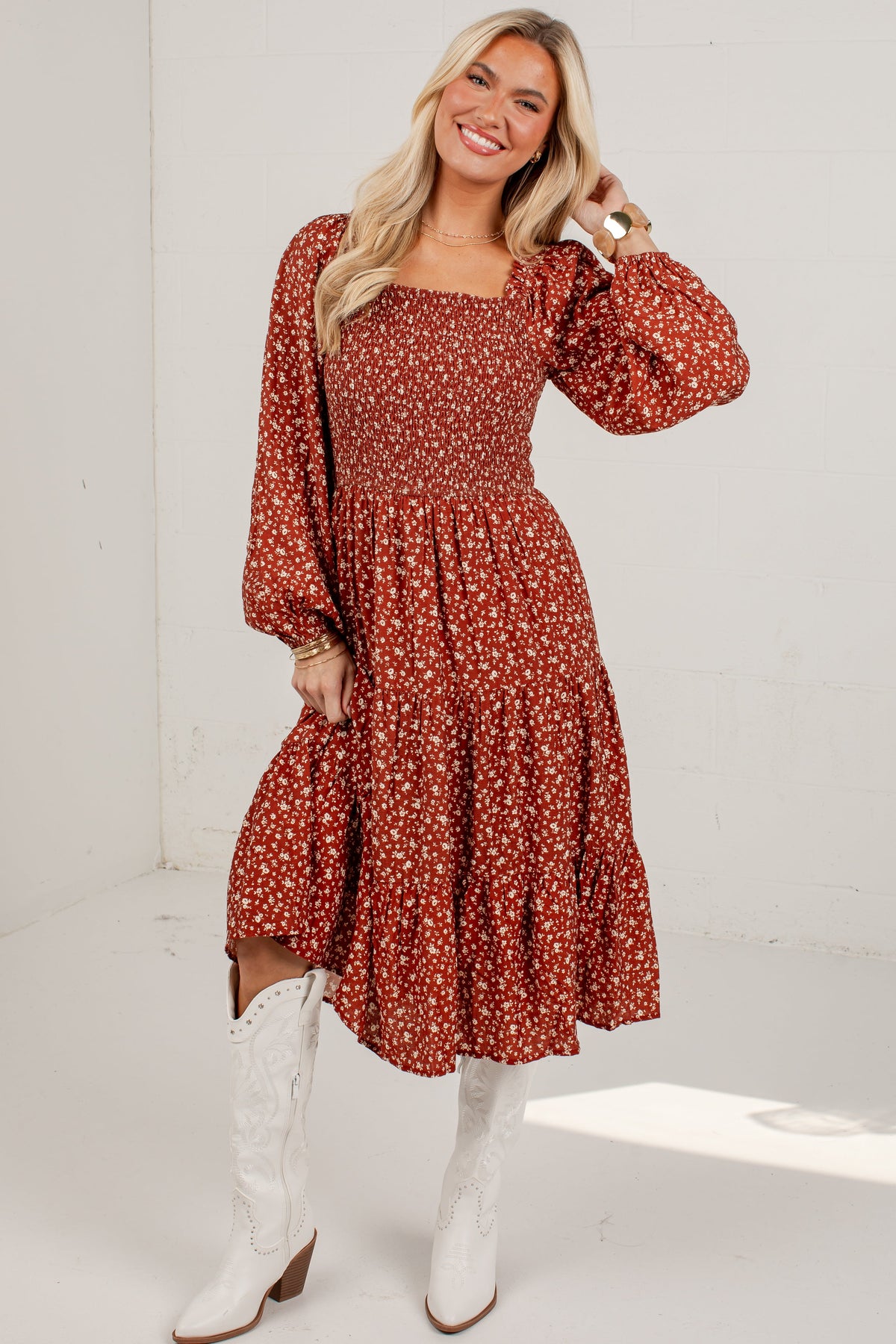 A woman modeling a rust-red long-sleeve midi dress with a white floral print, paired with white cowboy boots, standing against a white wall.