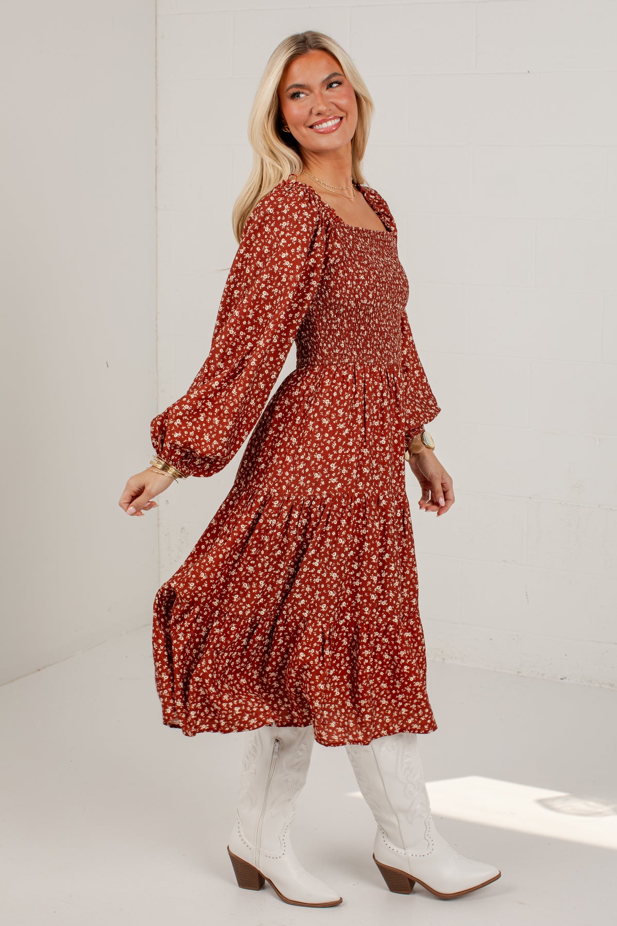 A woman modeling a rust-colored long-sleeve midi dress with a small white floral print, paired with white cowboy boots, in a bright white studio setting.