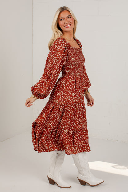A woman modeling a rust-colored long-sleeve midi dress with a small white floral print, paired with white cowboy boots, in a bright white studio setting.