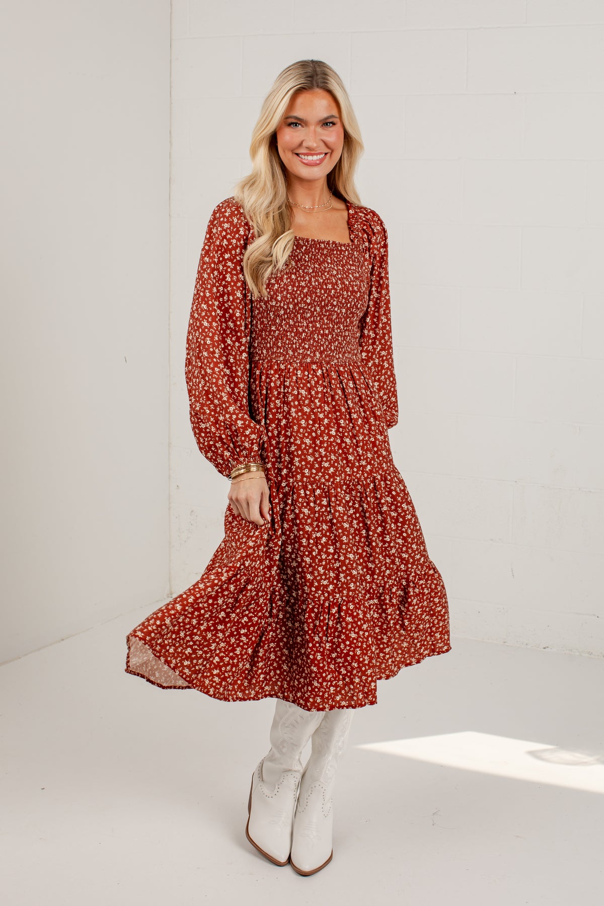 A woman modeling a rust-red long-sleeve midi dress with a white and orange floral pattern, paired with white knee-high boots, in a bright white studio setting.