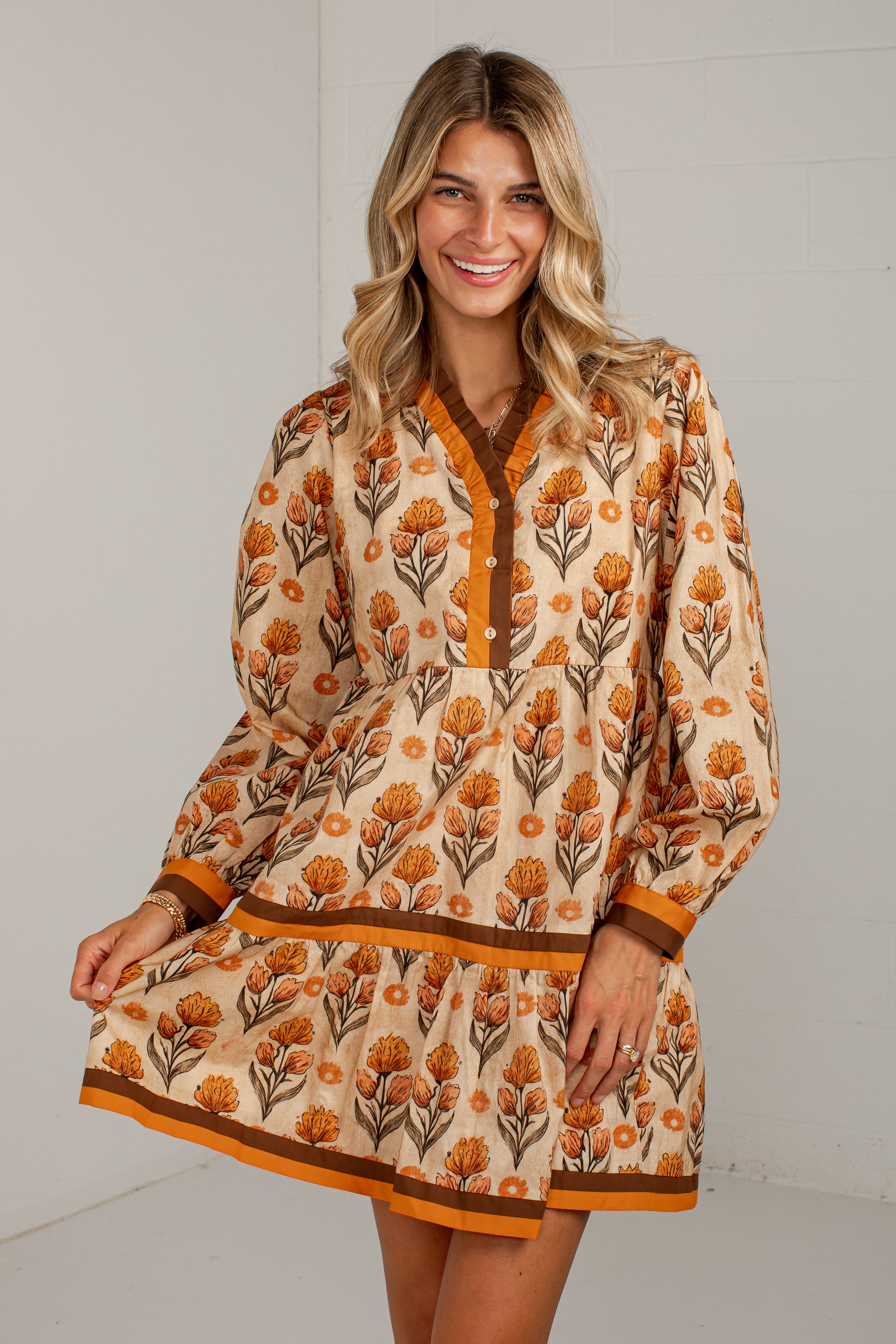 A woman modeling a long-sleeve tunic dress with a beige base, orange floral pattern, and brown trim against a light gray wall.