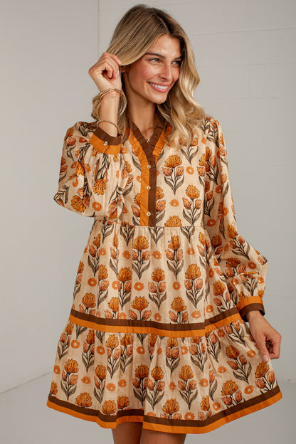 A woman wearing a tiered long-sleeve dress with a beige base, orange floral print, and brown trim, posing against a light gray wall.