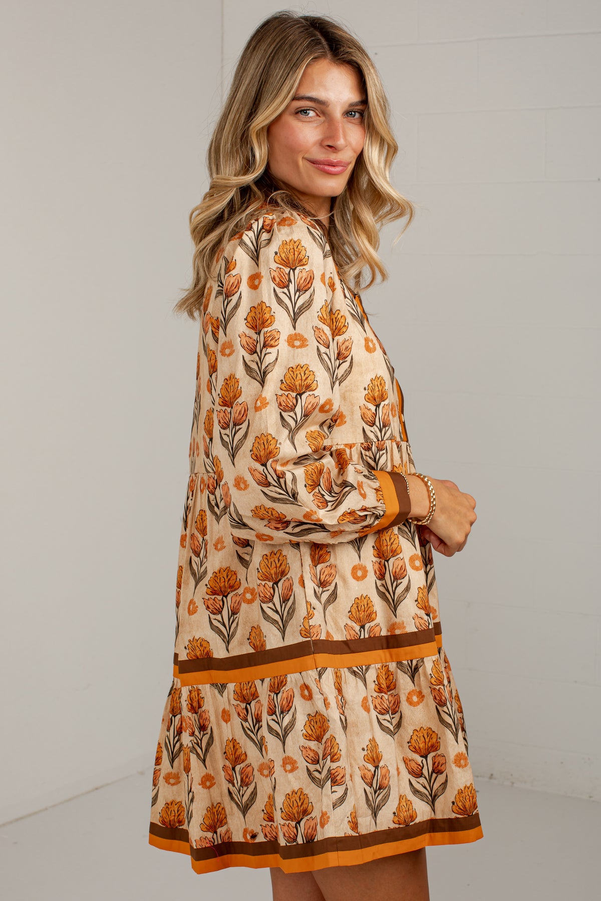 A woman wearing a long-sleeve tiered dress with an orange and brown floral pattern on a cream background, photographed against a light gray wall.