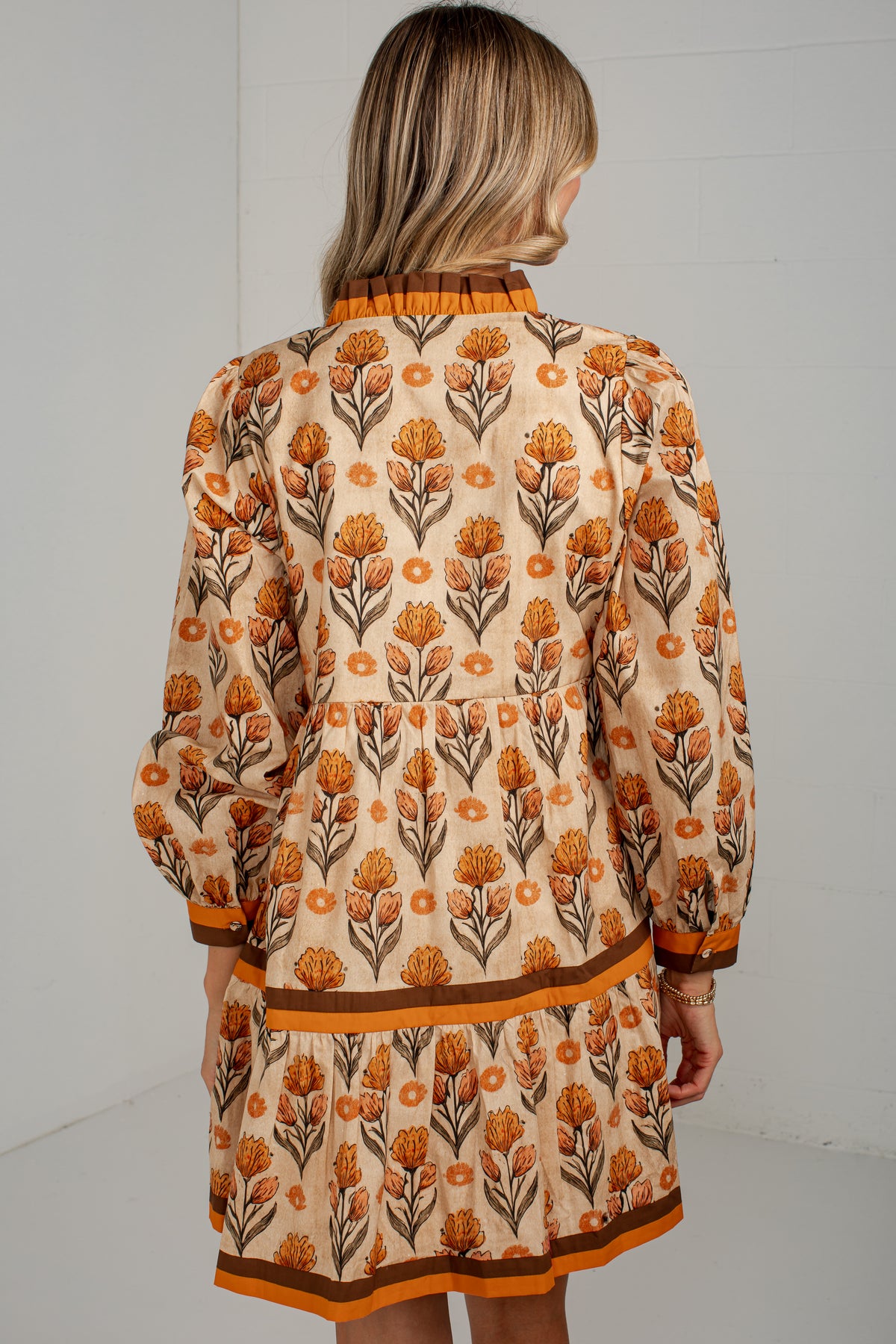 A woman viewed from the back wearing a beige long-sleeve mini dress with an orange and brown floral print and orange-brown trim.