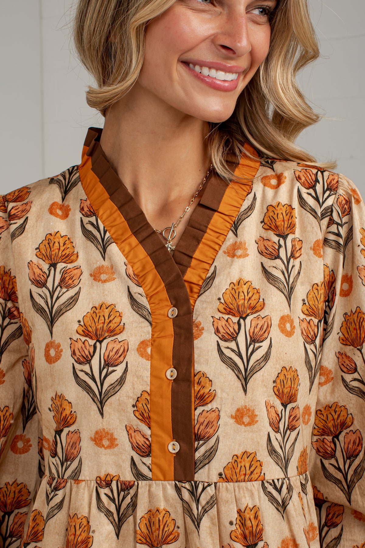 A close-up of a woman wearing a beige long-sleeve blouse with an orange and brown floral print, featuring a ruffled V-neckline and a brown button placket.