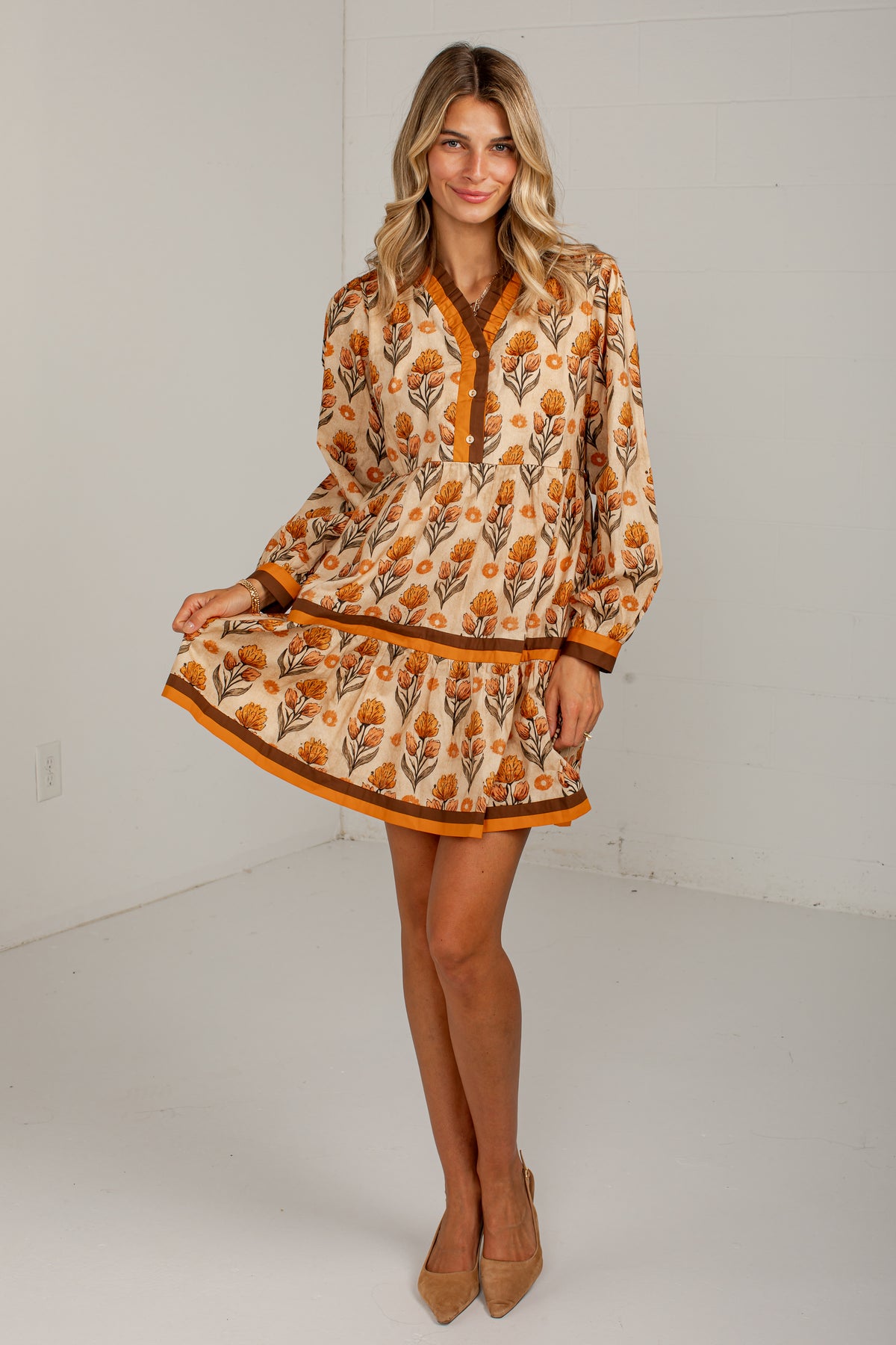 A woman modeling a long-sleeve mini dress with a warm-toned floral print in orange, brown, and beige on a cream background, paired with tan heels, set against a plain white studio background.