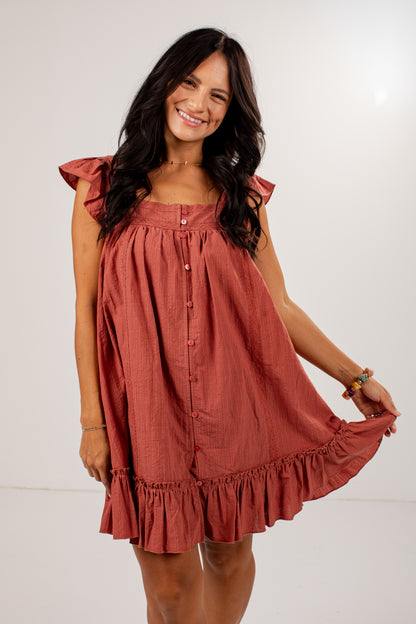 A woman modeling a rust-colored linen dress with ruffled sleeves and a button-down front, set against a plain white studio background.