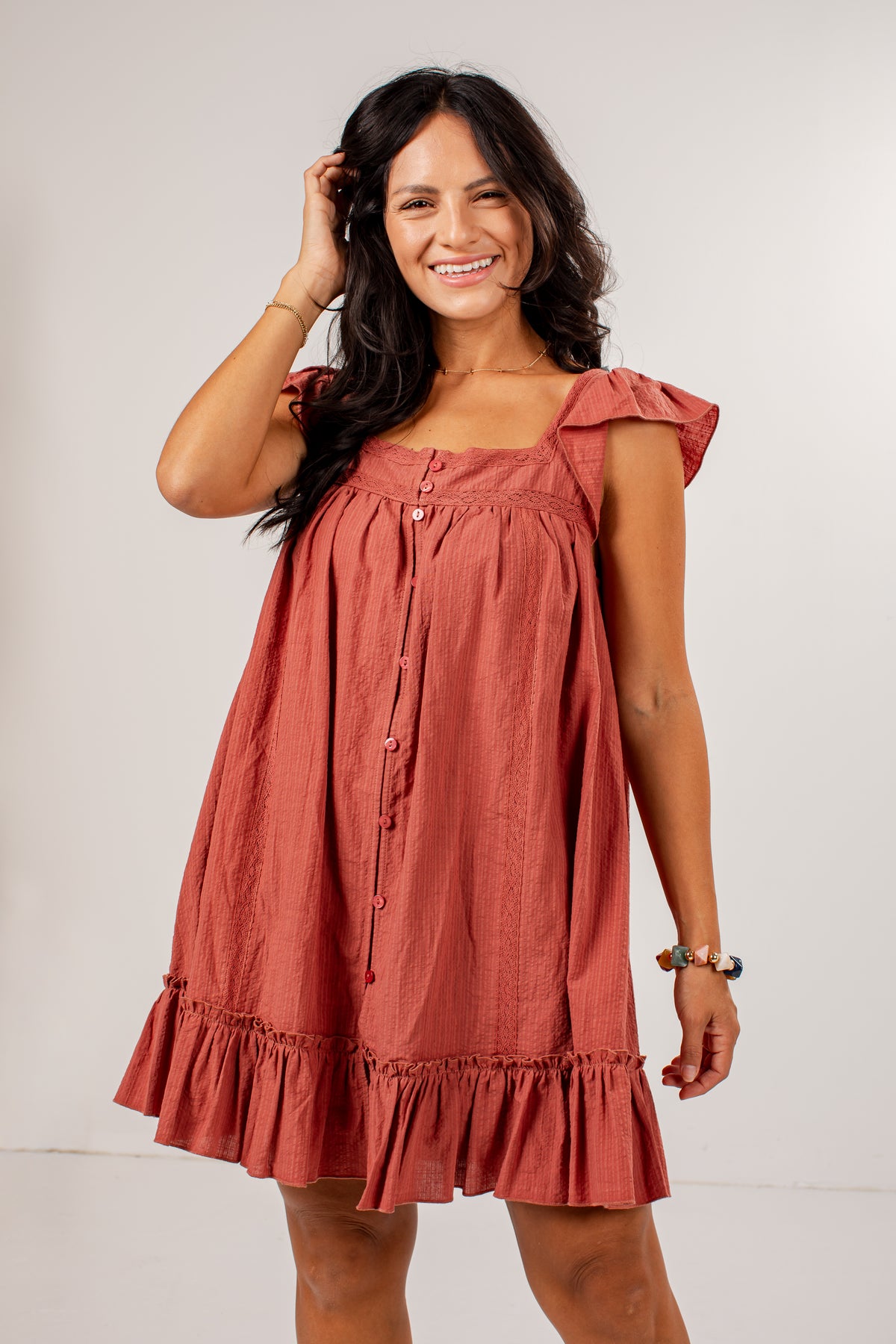 A woman modeling a terracotta-colored linen dress with ruffled shoulder straps, a button-down front, and a ruffled hem against a plain light gray background.