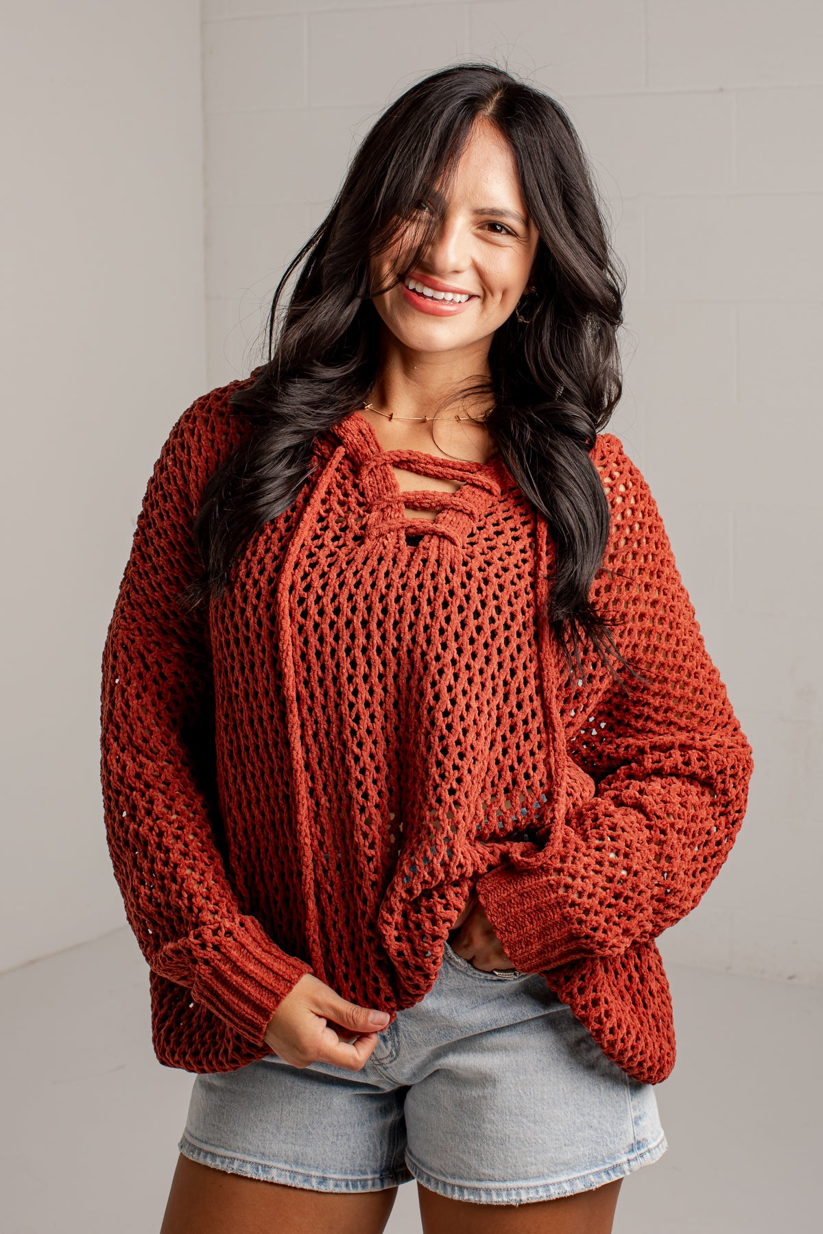 A woman wearing a rust-orange chunky knit lace-up sweater and light-wash denim shorts, standing against a light gray wall.