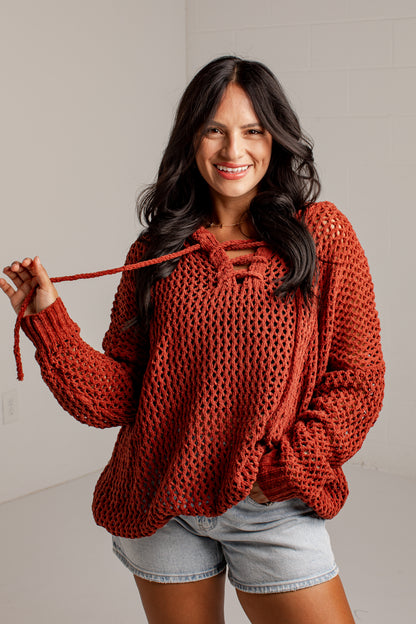 A woman wearing a chunky, open-knit rust-orange sweater with a lace-up neckline and denim shorts, holding up the tie detail of the sweater against a light gray wall.