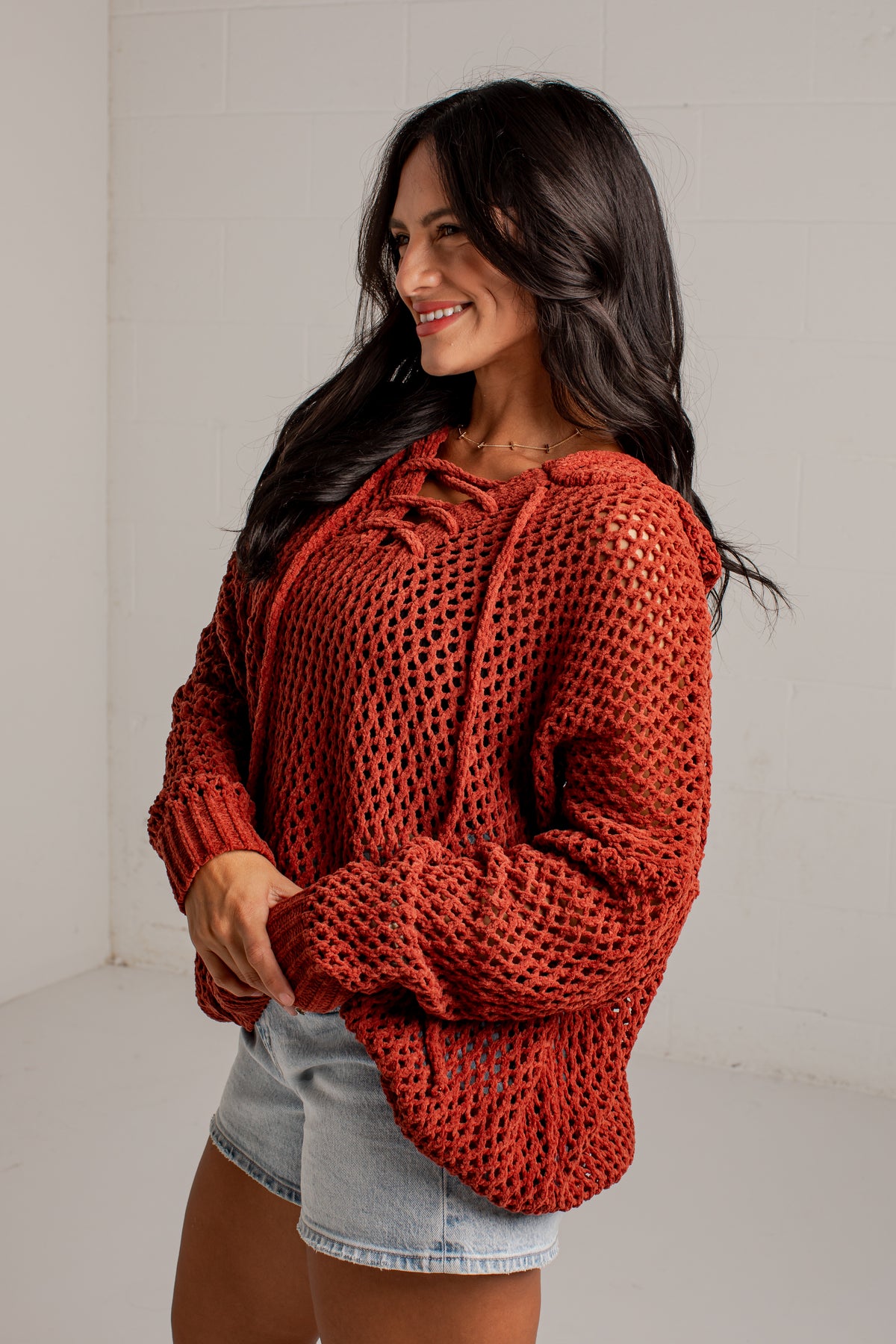 A woman wearing a rust-orange chunky open-knit pullover sweater with a lace-up front and light-wash denim shorts, standing against a white wall.