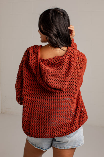 A woman viewed from behind wearing a rust-orange open-knit hooded sweater and light-wash denim shorts against a plain light gray wall.