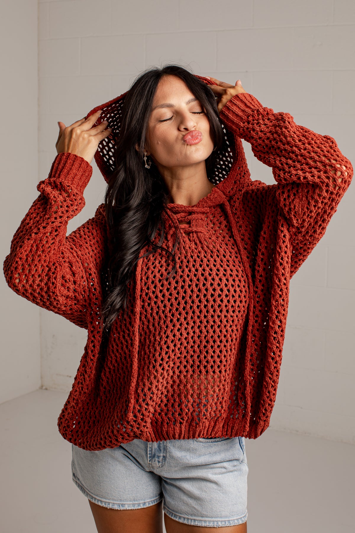 A woman wearing a chunky, open-knit rust-orange hoodie with drawstrings, posing with her hands on her hood in a bright studio setting.