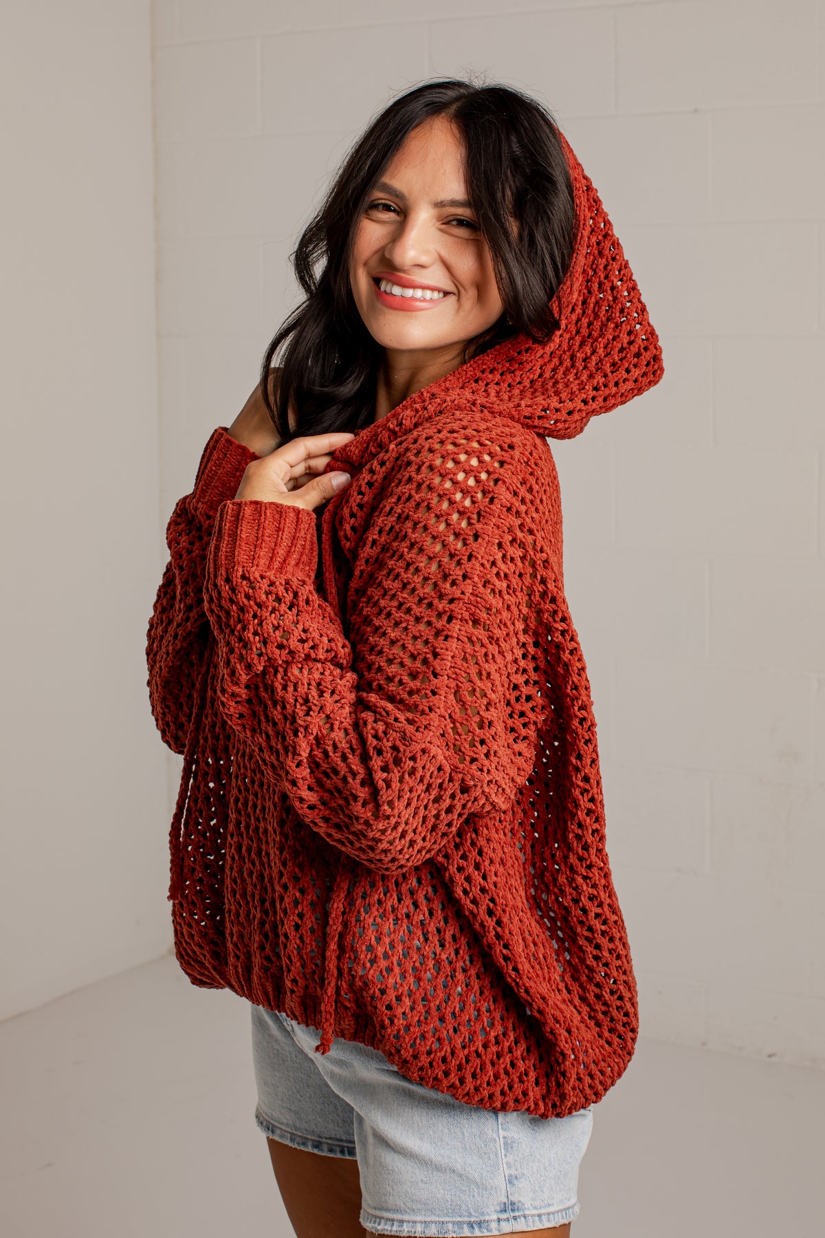 A woman wearing a rust-orange chunky open-knit hooded sweater and light-wash denim shorts, standing against a light gray wall.