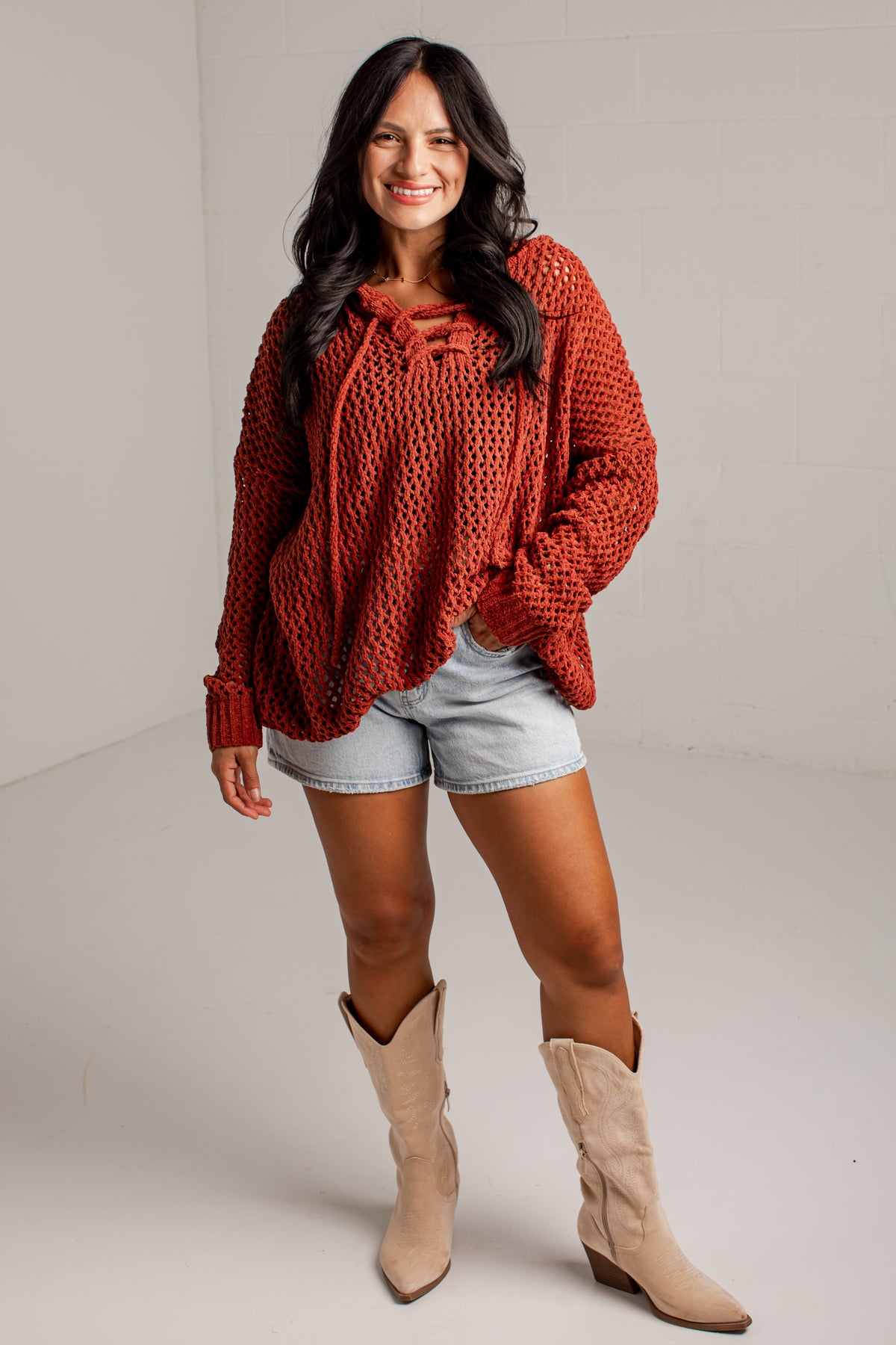 A woman modeling a rust-orange chunky knit long-sleeve top with tie details at the neckline, paired with light-wash denim shorts and beige cowboy boots, against a plain light gray studio background.