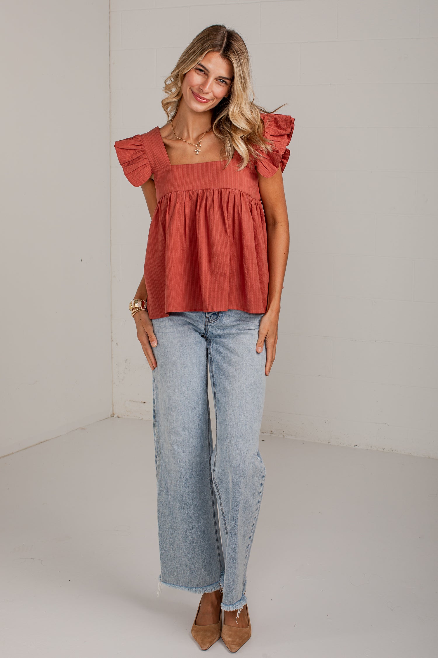 A woman modeling a terracotta-colored ruffled off-the-shoulder top paired with light-wash wide-leg jeans and brown pointed-toe heels, standing against a white studio wall.