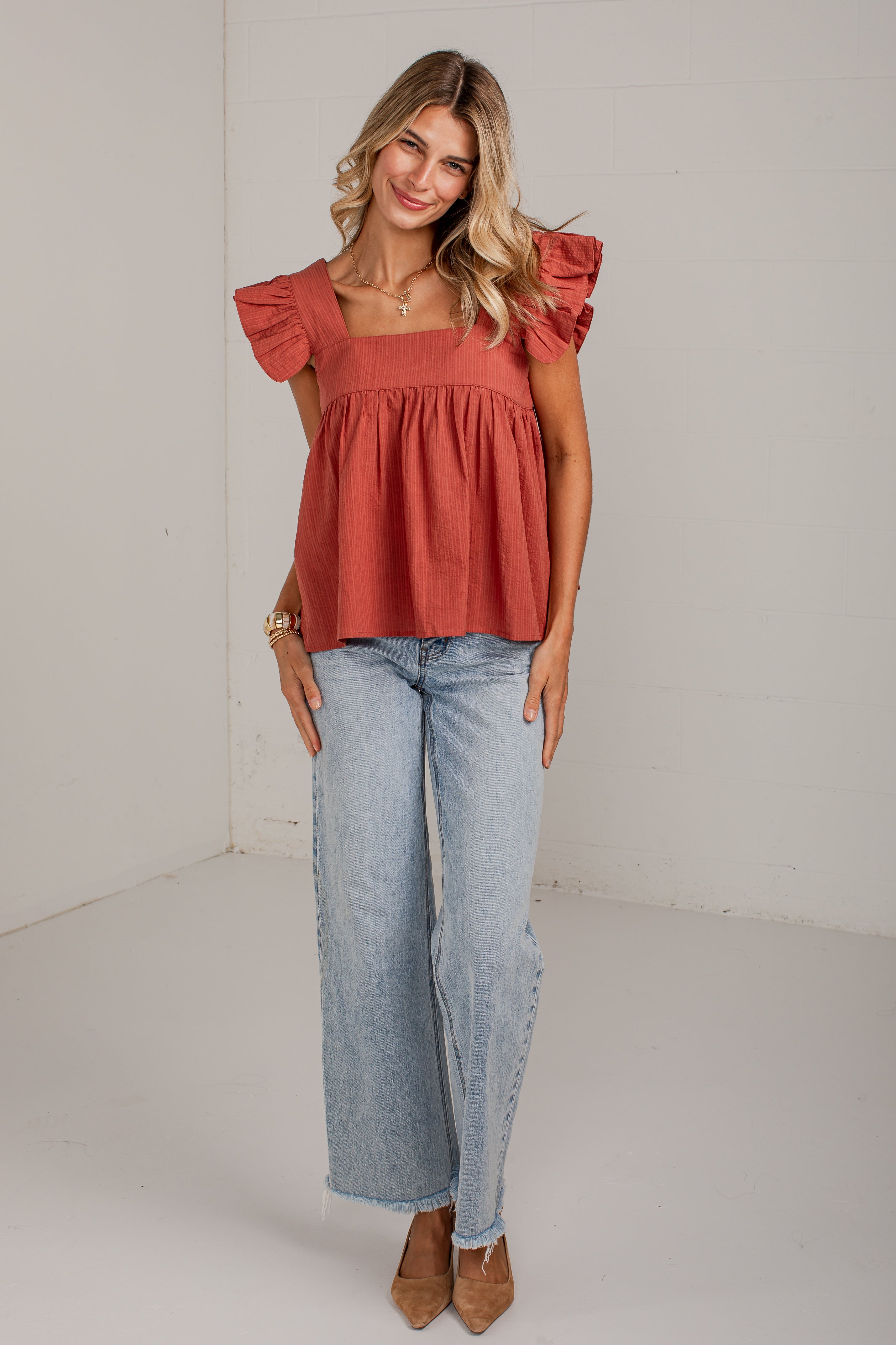 A woman modeling a terracotta-colored ruffled off-the-shoulder top paired with light-wash wide-leg jeans and brown pointed-toe heels, standing against a white studio wall.