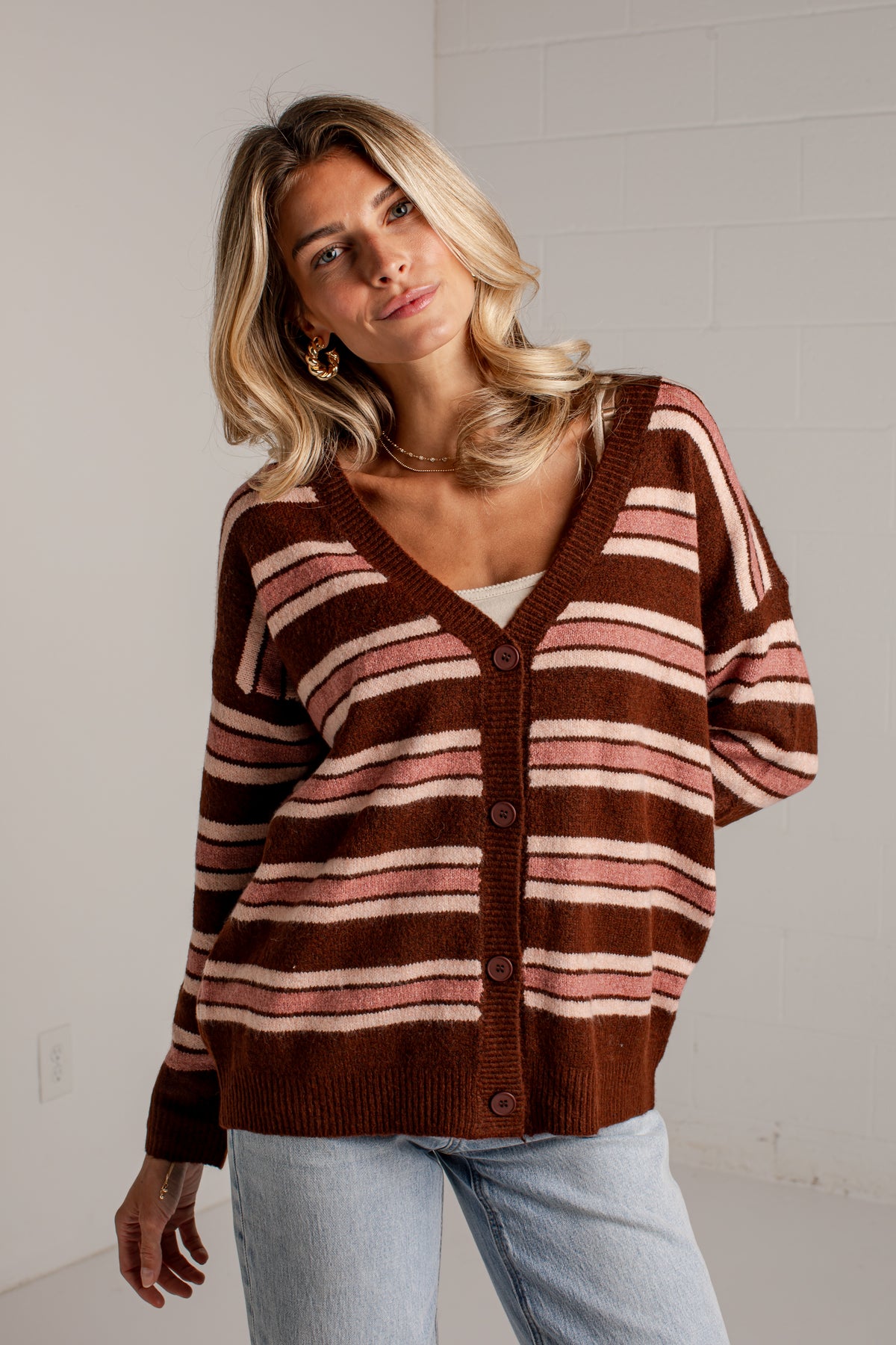 A woman wearing a brown and pink striped knit cardigan sweater with light blue jeans, standing against a white brick wall.