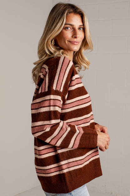 A woman wearing a brown knit sweater with horizontal stripes in dusty pink and cream, photographed against a light gray brick wall.