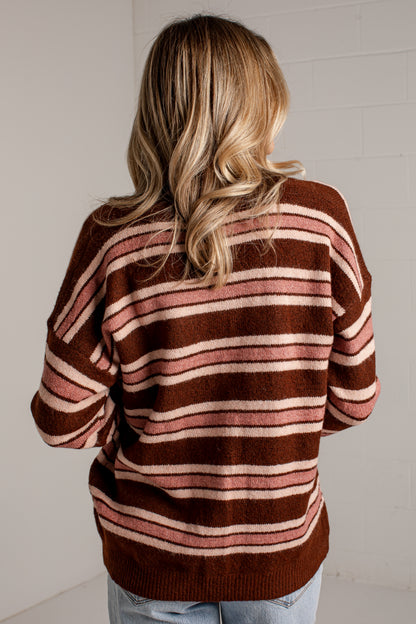 A woman viewed from behind wearing a brown knit sweater with horizontal pink and cream stripes, paired with light blue jeans, standing against a white brick wall.