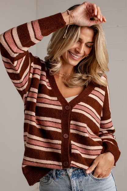 A woman wearing a brown, pink, and cream striped knit cardigan sweater with button-front closure, paired with light blue jeans, posing against a light gray wall.