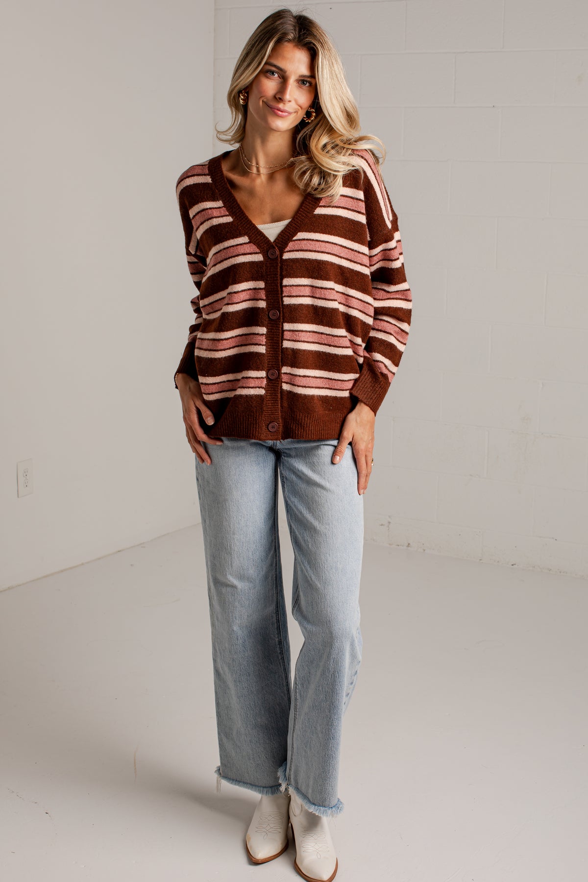 A woman modeling a brown and beige striped cardigan sweater, paired with light-wash jeans and white boots, standing in a bright studio setting.