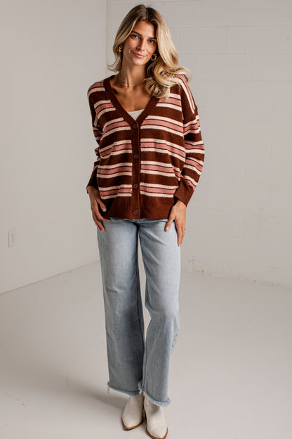 A woman modeling a brown and beige striped cardigan sweater, paired with light-wash jeans and white boots, standing in a bright studio setting.
