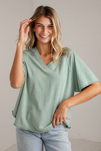 A woman wearing a sage green short-sleeve V-neck top and light blue jeans, standing against a light gray wall.