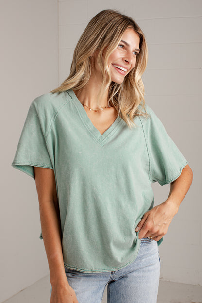 A woman wearing a light sage green V-neck short-sleeve top and light wash blue jeans, standing against a light gray paneled wall.