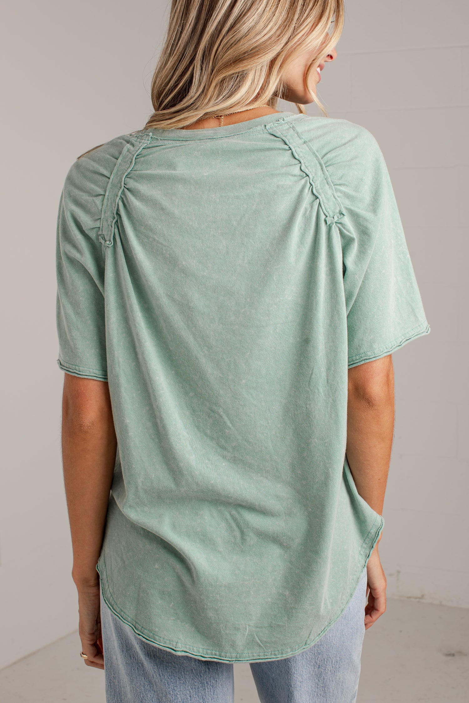 A woman shown from the back wearing a sage green short-sleeve top with gathered shoulder details and a curved hem, paired with light blue jeans.