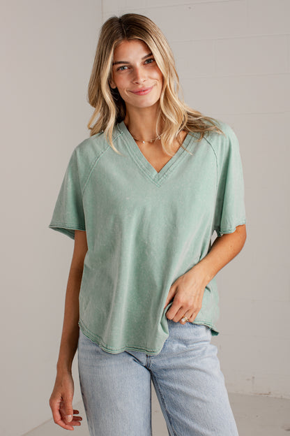 A woman wearing a sage green short-sleeve V-neck top and light blue jeans, standing against a light gray wall.