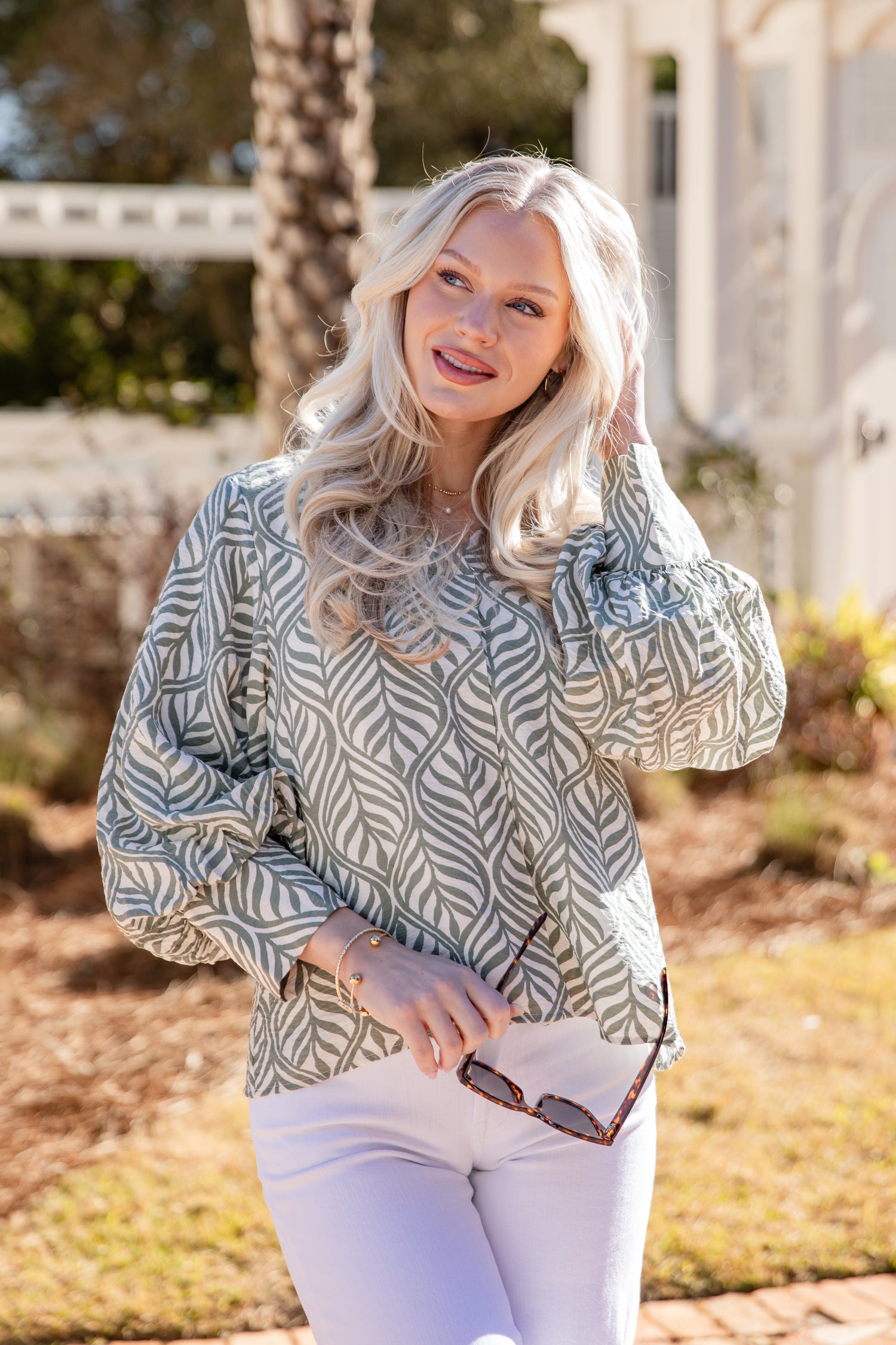 Woman wearing a patterned blouse and white pants outdoors.