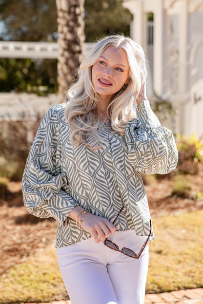 Woman wearing a patterned blouse and white pants outdoors.